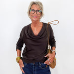 A woman with short gray hair and glasses wearing a dark brown cowl-neck top and blue jeans, holding a tan shoulder bag against a plain white background.