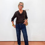 A woman modeling a dark brown cowl-neck long-sleeve top, blue jeans, and beige loafers, carrying a tan shoulder bag, standing against a plain white wall.