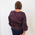 Person wearing a patterned blouse and dark pants against a white wall
