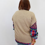 A woman viewed from behind wearing a beige knit sweater with colorful patterned fabric panels on the sleeves and hem, paired with blue jeans against a plain white background.