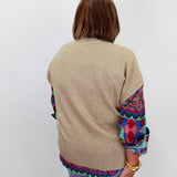 A woman viewed from behind wearing a beige knit sweater with colorful patterned fabric panels on the sleeves and hem, paired with blue jeans against a plain white background.
