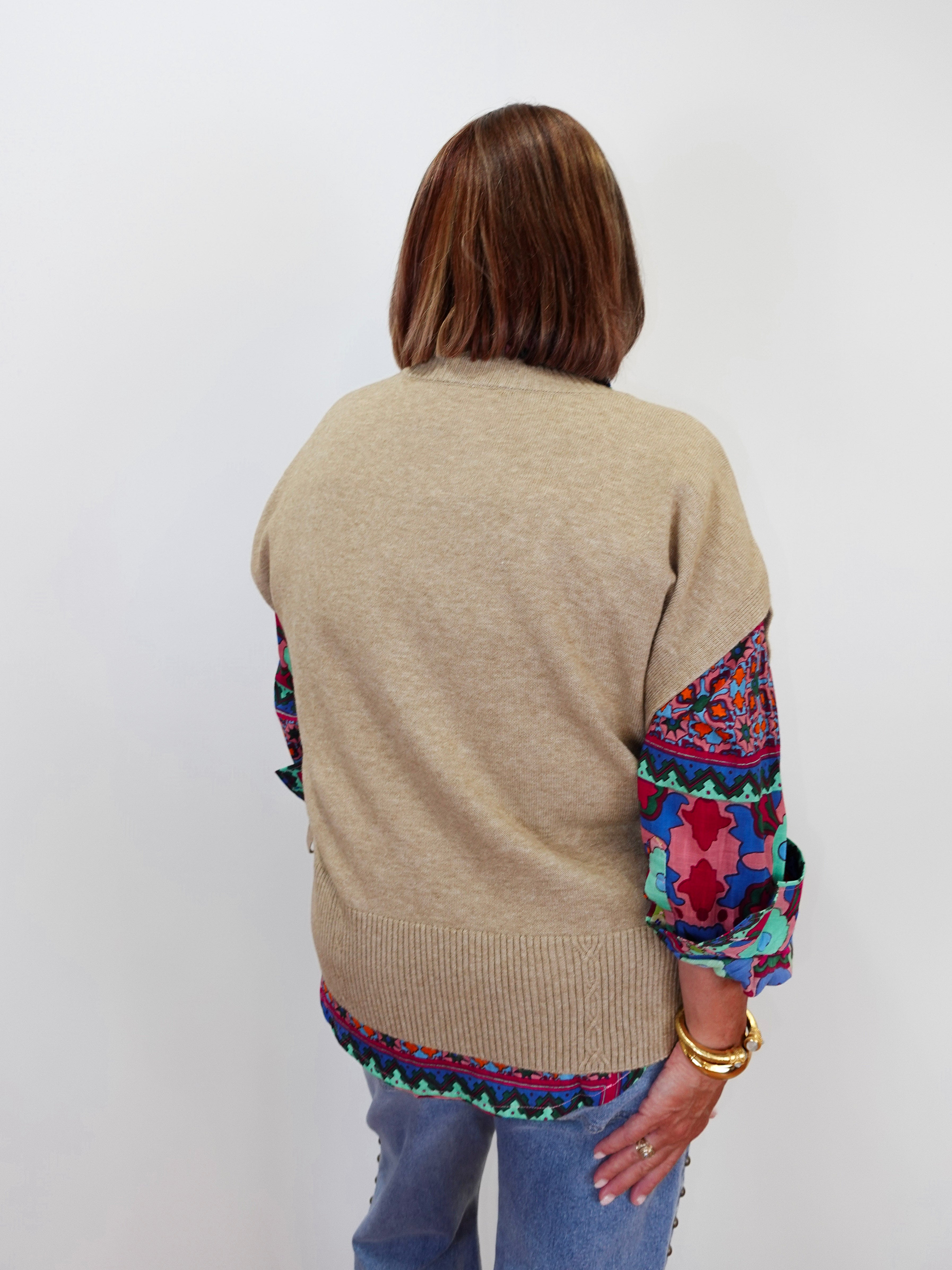 A woman viewed from behind wearing a beige knit sweater with colorful patterned fabric panels on the sleeves and hem, paired with blue jeans against a plain white background.