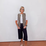 Woman wearing a patterned vest, white shirt, black pants, and white shoes against a plain white wall.