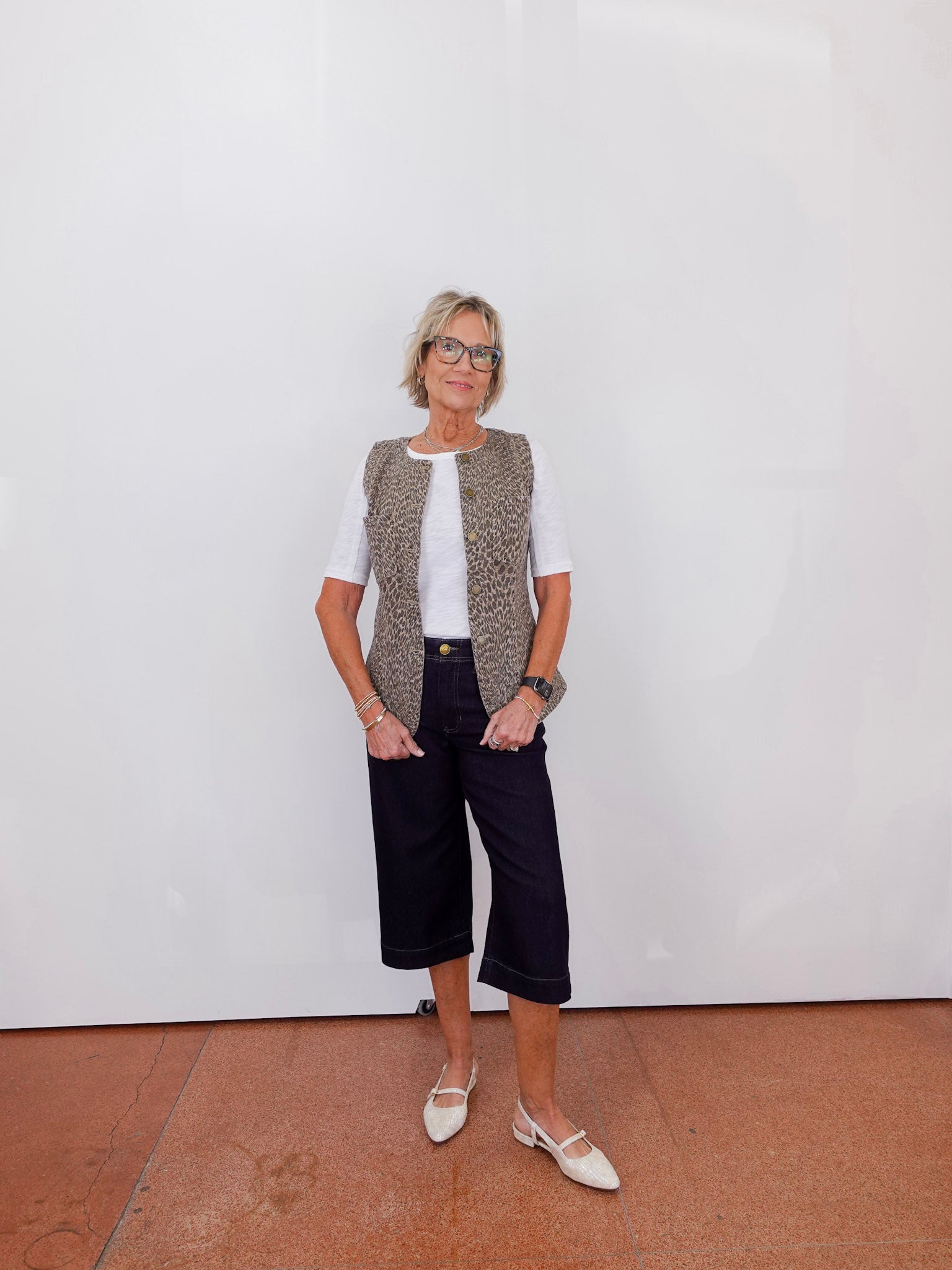 Woman wearing a patterned vest, white shirt, black pants, and white shoes against a plain white wall.