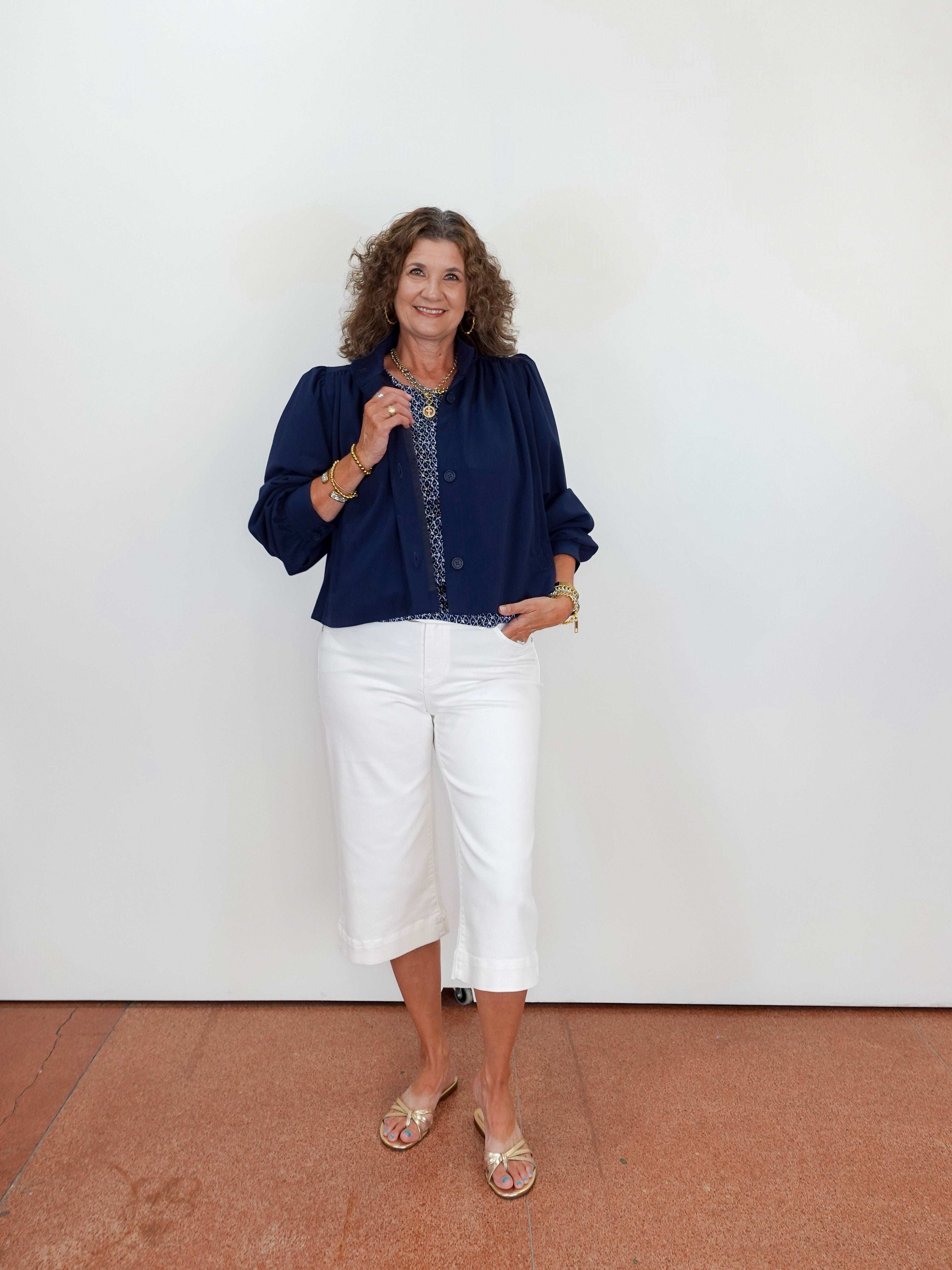 Woman wearing a navy blue top and white pants standing against a plain white wall.