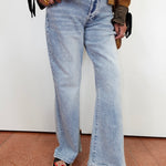 A person wearing light-wash wide-leg denim jeans, styled with a brown jacket, striped top, and black slide sandals with red nail polish, standing against a plain white wall.