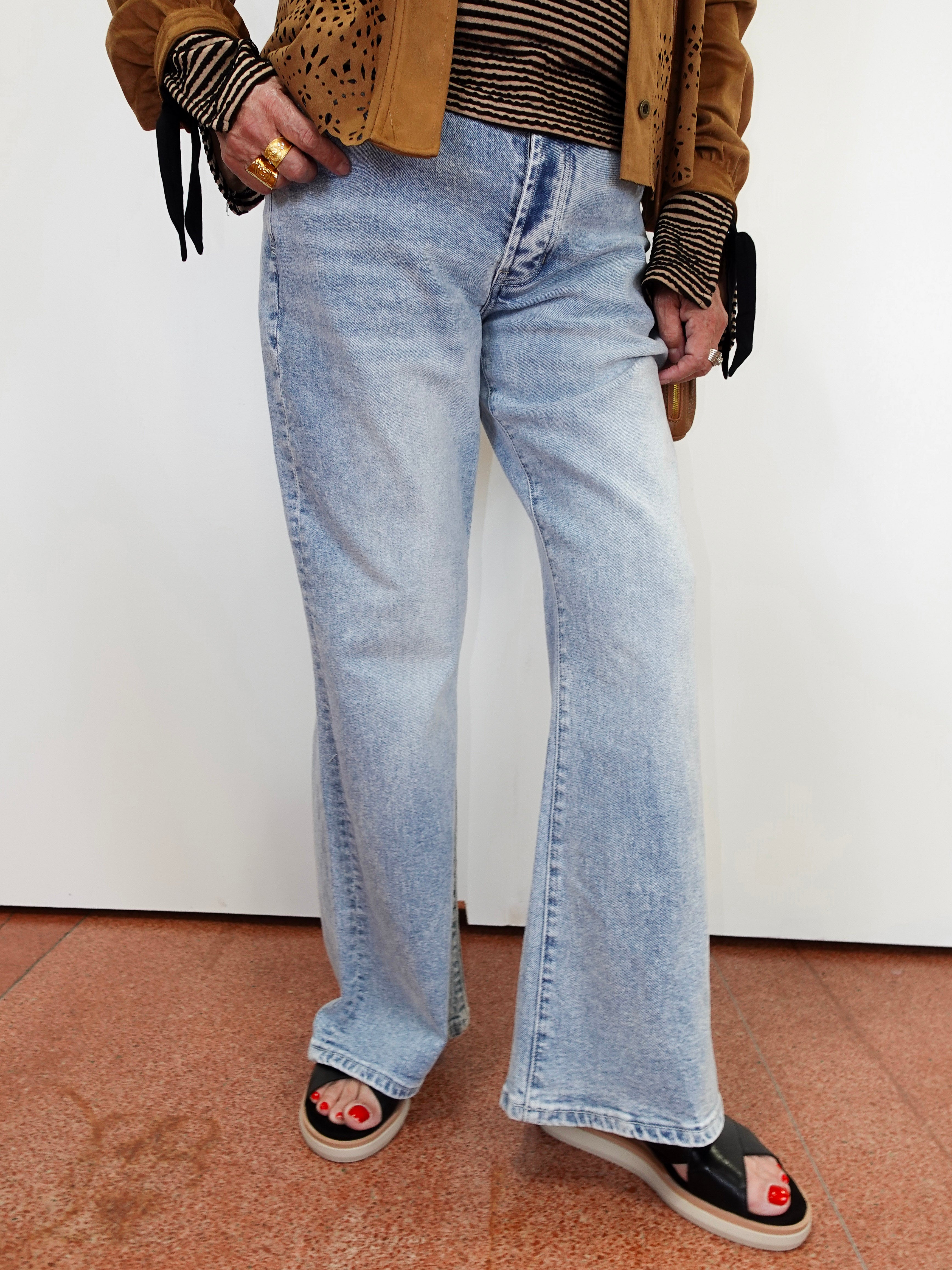 A person wearing light-wash wide-leg denim jeans, styled with a brown jacket, striped top, and black slide sandals with red nail polish, standing against a plain white wall.