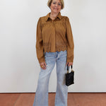 A woman stands against a plain white wall wearing a brown long-sleeve blouse with lace-up detailing at the hem, light-wash wide-leg jeans, black slide sandals, and holding a small black handbag.