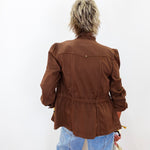 A person viewed from behind wearing a brown cropped jacket with puffed shoulders and a gathered waist, paired with light blue jeans against a plain white background.