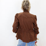 A person viewed from behind wearing a brown cropped jacket with puffed shoulders and a gathered waist, paired with light blue jeans against a plain white background.