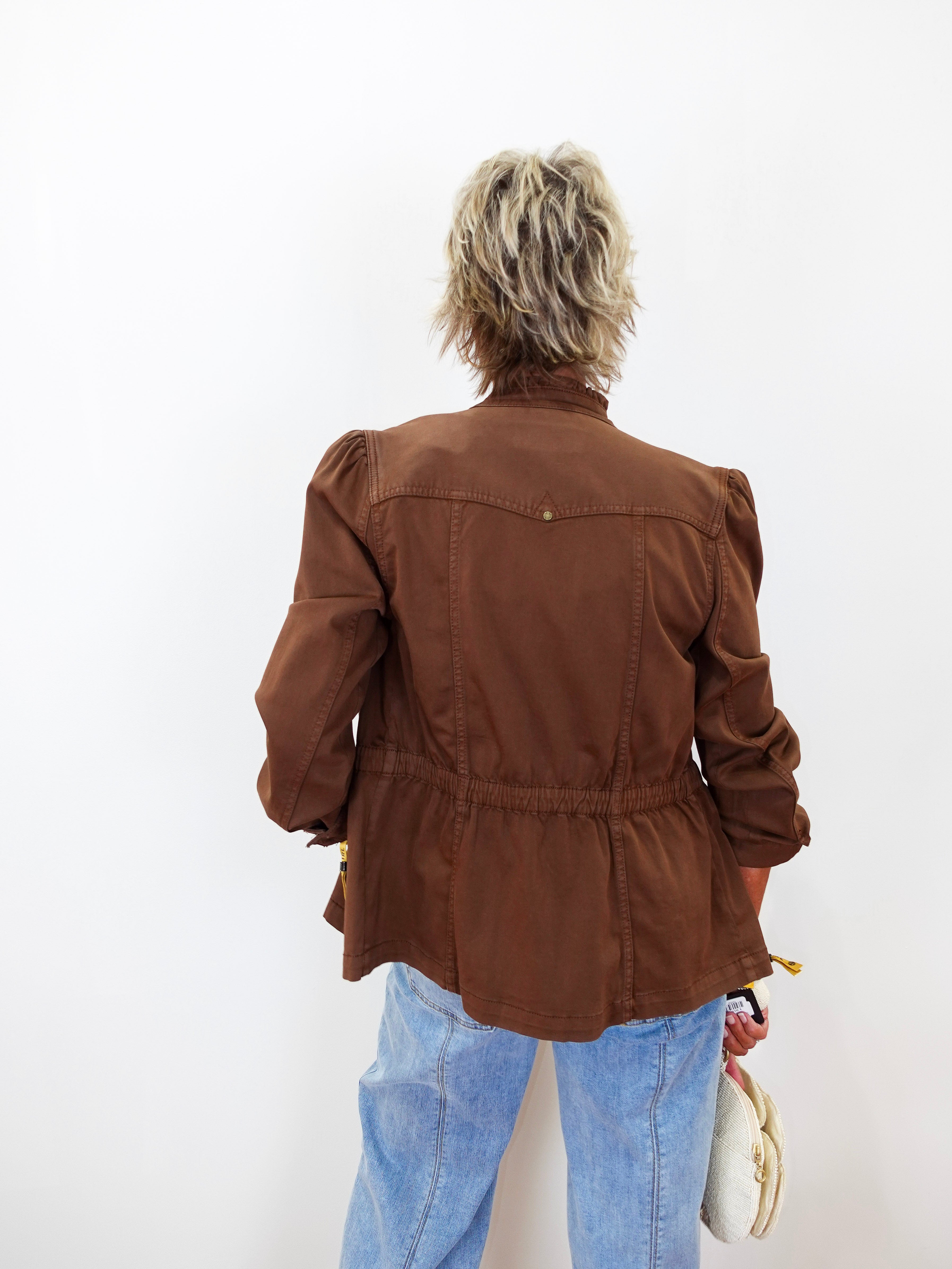 A person viewed from behind wearing a brown cropped jacket with puffed shoulders and a gathered waist, paired with light blue jeans against a plain white background.
