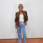 A woman stands against a white wall wearing a brown jacket over a white top, light blue wide-leg jeans, and beige sandals, holding a small cream clutch.