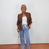 A woman stands against a white wall wearing a brown jacket over a white top, light blue wide-leg jeans, and beige sandals, holding a small cream clutch.