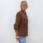 A woman with short blonde hair and glasses, wearing a brown utility jacket and light blue jeans, holding a white clutch bag against a plain white background.