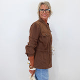 A woman with short blonde hair and glasses, wearing a brown utility jacket and light blue jeans, holding a white clutch bag against a plain white background.