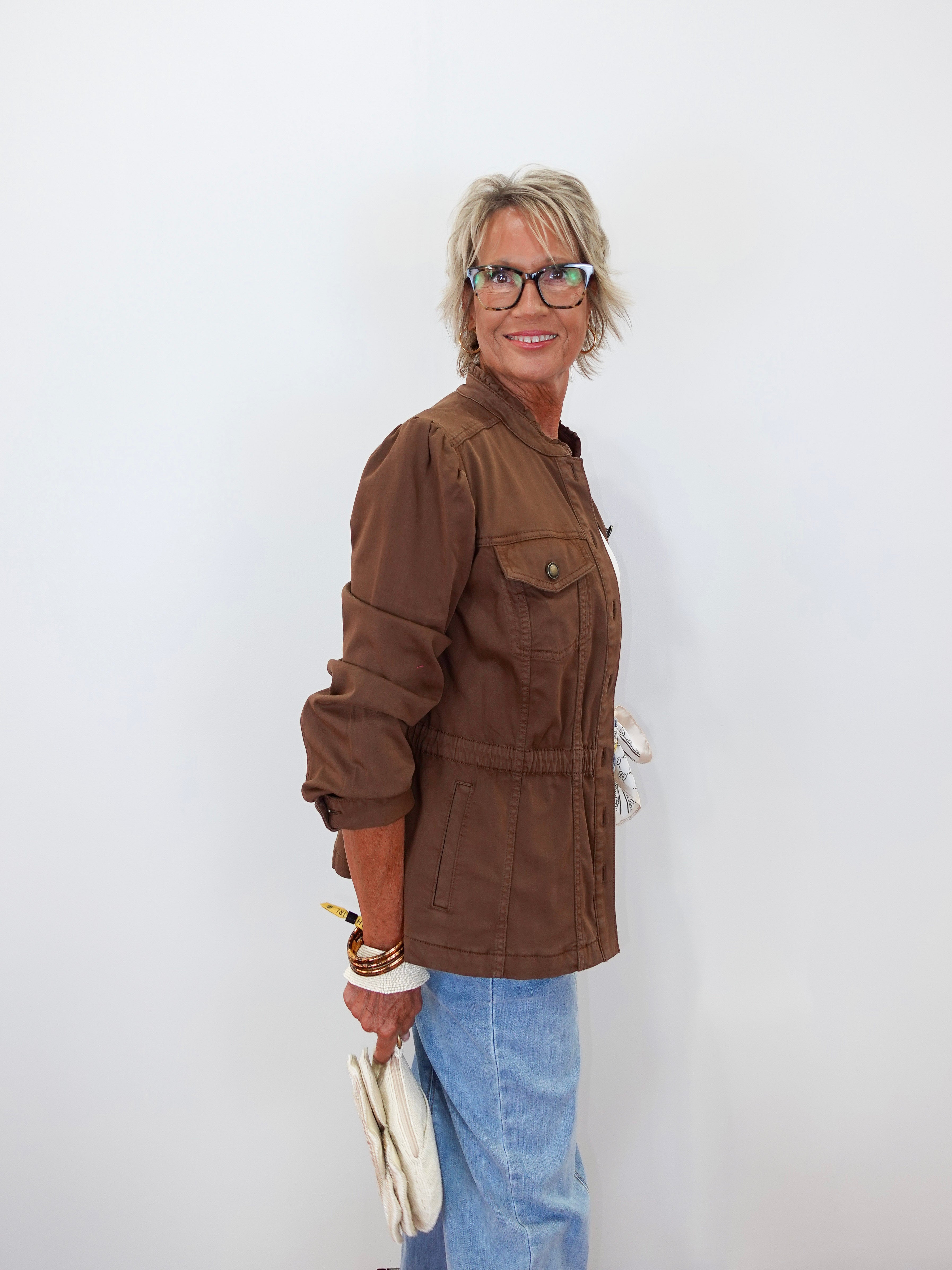 A woman with short blonde hair and glasses, wearing a brown utility jacket and light blue jeans, holding a white clutch bag against a plain white background.