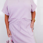 Person wearing a light purple outfit with a matching handbag on a plain background