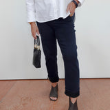 A person wearing a white button-up shirt, dark navy cropped pants, and dark textured peep-toe heels, holding a small black handbag against a plain white wall and terracotta floor.