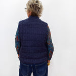 A person viewed from the back wearing a navy blue cable-knit quilted vest over a multicolored knit long-sleeve shirt and blue jeans, standing against a plain white wall.