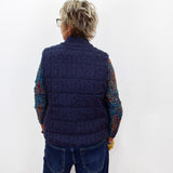A person viewed from the back wearing a navy blue cable-knit quilted vest over a multicolored knit long-sleeve shirt and blue jeans, standing against a plain white wall.