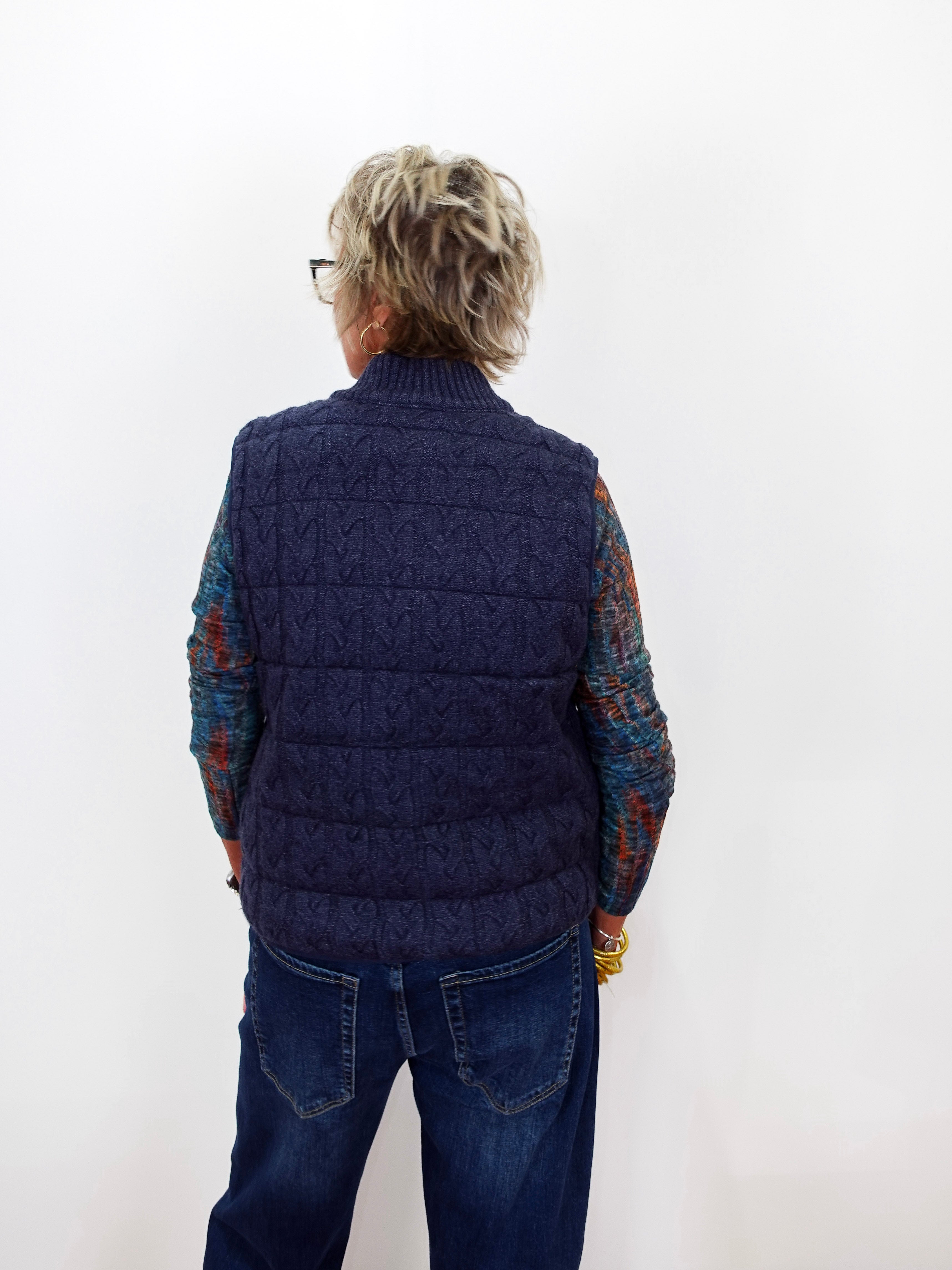 A person viewed from the back wearing a navy blue cable-knit quilted vest over a multicolored knit long-sleeve shirt and blue jeans, standing against a plain white wall.