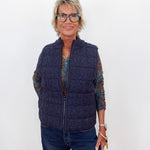 A woman with short blonde hair and glasses wearing a navy blue cable-knit zip-up puffer vest over a patterned long-sleeve shirt and blue jeans, standing against a plain white background.