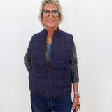 A woman with short blonde hair and glasses wearing a navy blue cable-knit zip-up puffer vest over a patterned long-sleeve shirt and blue jeans, standing against a plain white background.