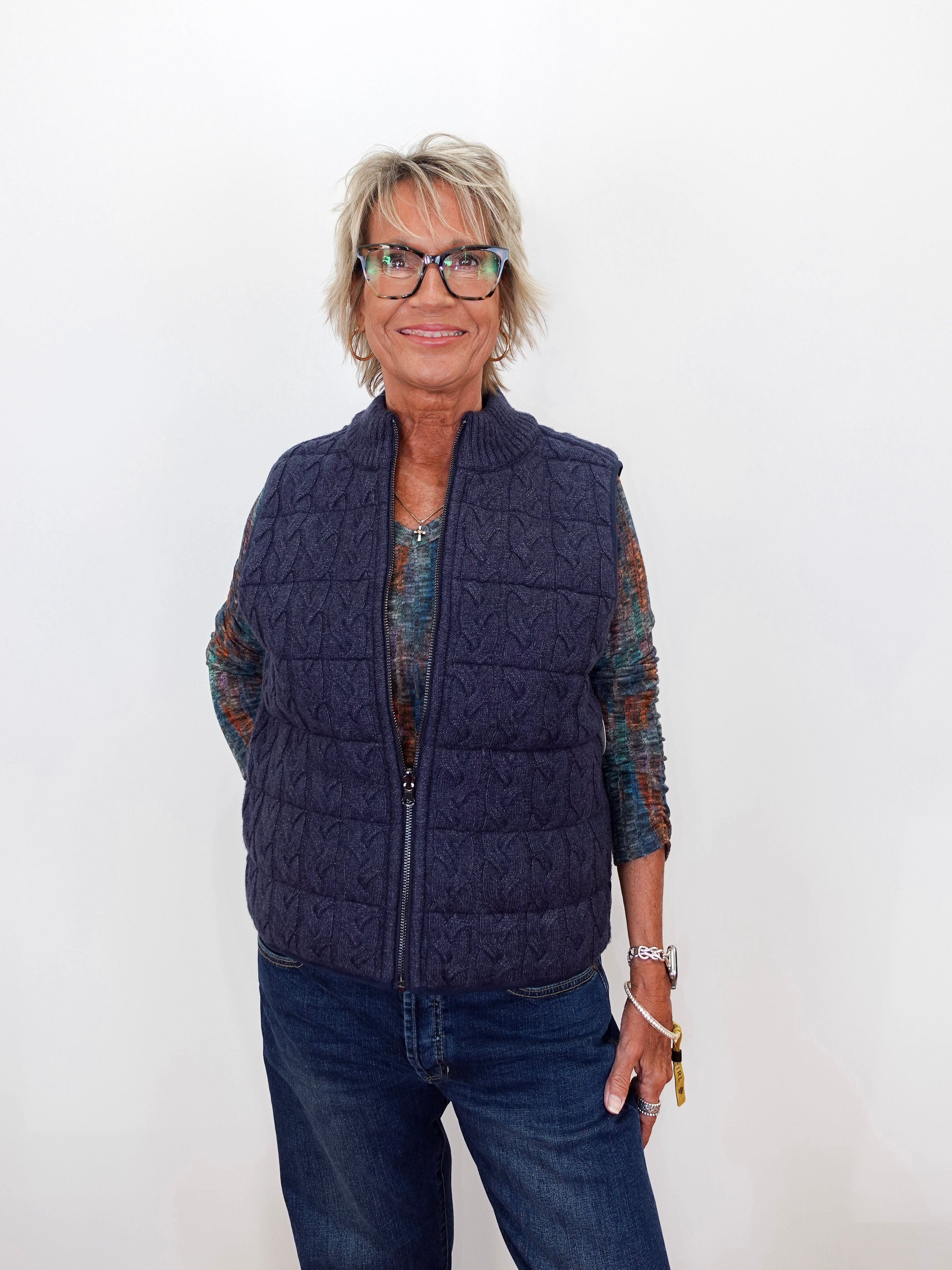 A woman with short blonde hair and glasses wearing a navy blue cable-knit zip-up puffer vest over a patterned long-sleeve shirt and blue jeans, standing against a plain white background.