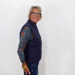A woman with short gray hair and glasses, wearing a navy blue cable-knit quilted vest over a multicolor knit long-sleeve shirt and blue jeans, standing against a plain white background.