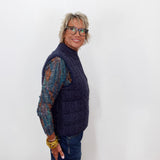 A woman with short gray hair and glasses, wearing a navy blue cable-knit quilted vest over a multicolor knit long-sleeve shirt and blue jeans, standing against a plain white background.
