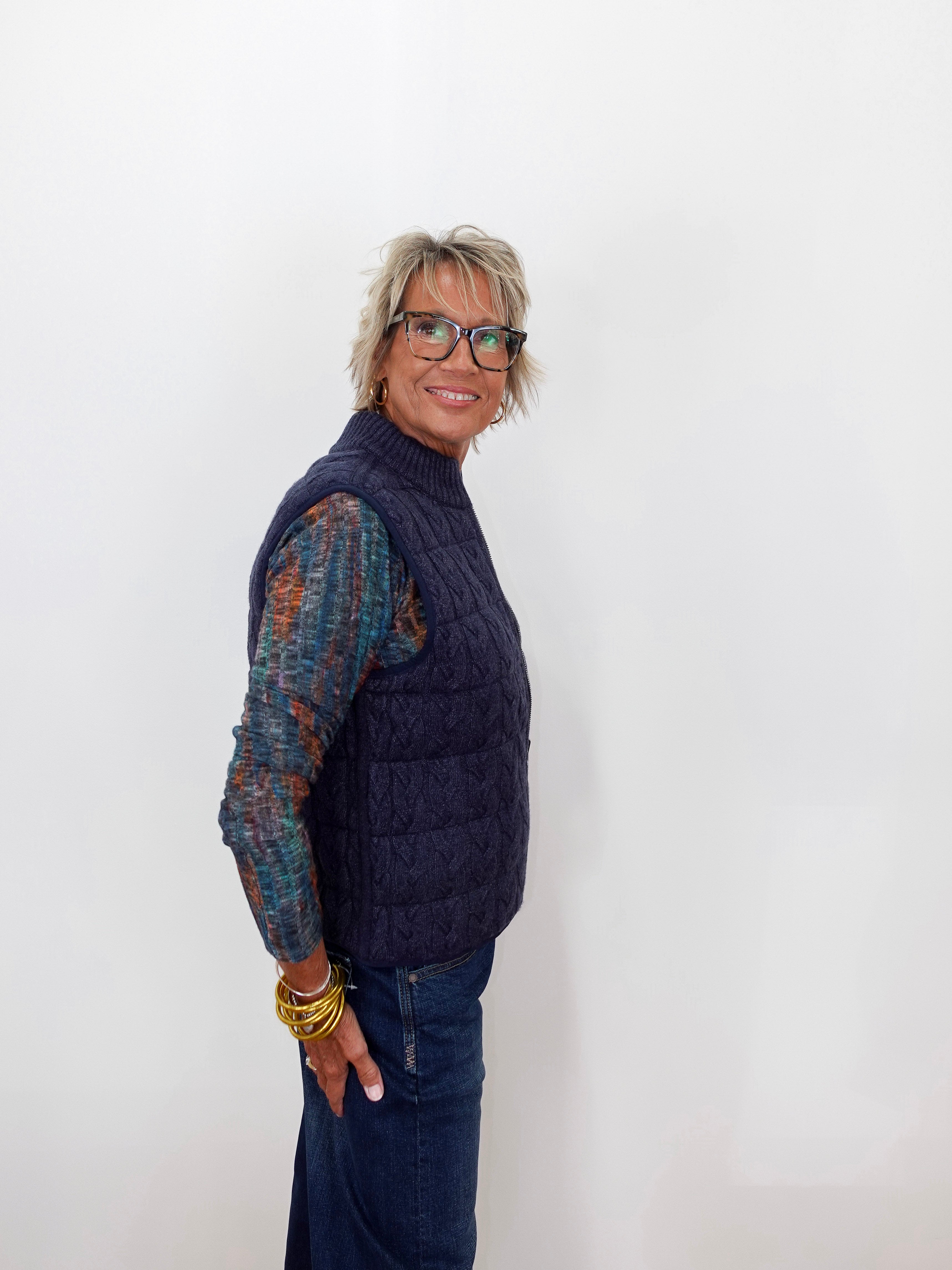 A woman with short gray hair and glasses, wearing a navy blue cable-knit quilted vest over a multicolor knit long-sleeve shirt and blue jeans, standing against a plain white background.