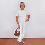 Woman in a white outfit holding a brown handbag against a white wall.