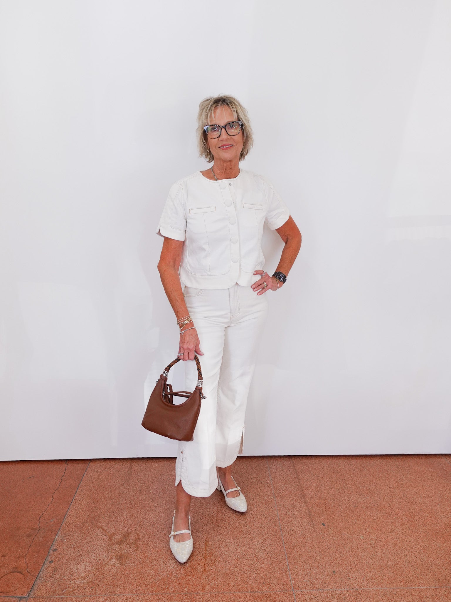 Woman in a white outfit holding a brown handbag against a white wall.