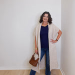 Woman wearing a long beige cardigan, blue jeans, and white sneakers in a room with a white wall.