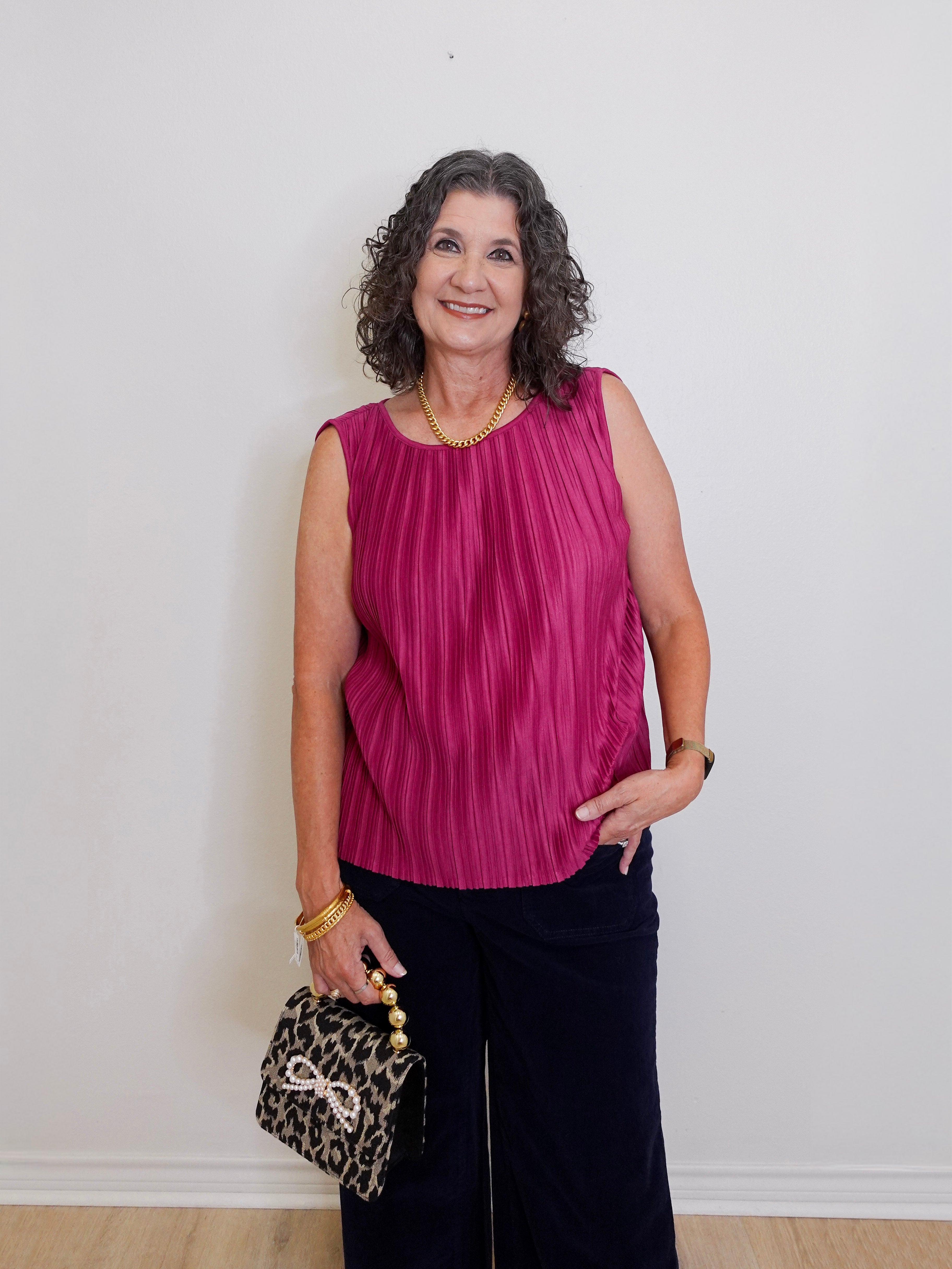 Woman wearing a pink sleeveless top and black pants, holding a leopard print handbag against a white wall.