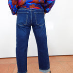 A person shown from the back wearing dark blue straight-leg jeans with a cuffed hem, paired with a colorful patterned blouse and black shoes, standing against a white wall.