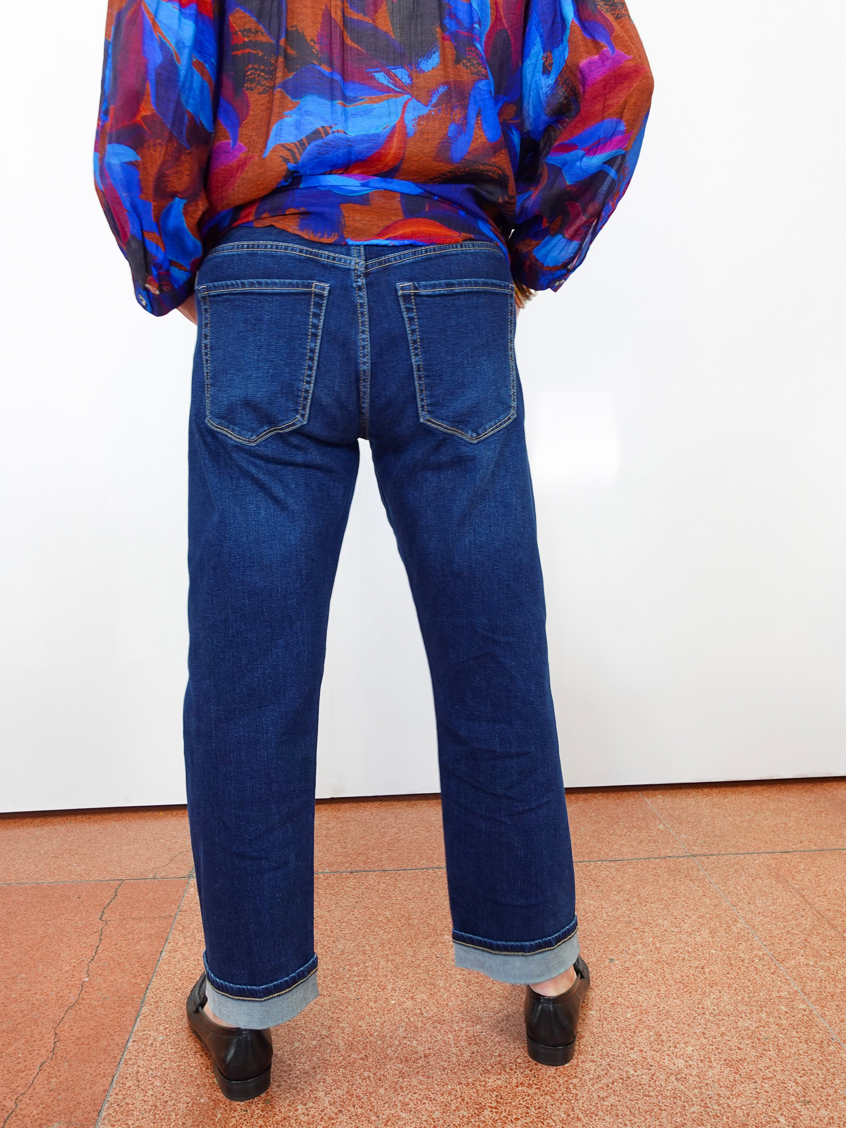 A person shown from the back wearing dark blue straight-leg jeans with a cuffed hem, paired with a colorful patterned blouse and black shoes, standing against a white wall.