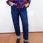 A person wearing dark blue straight-leg jeans with rolled cuffs, paired with a colorful abstract print button-up shirt and black loafers, standing against a white wall.