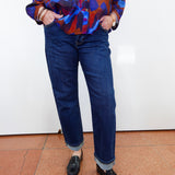 A person wearing dark blue straight-leg jeans with rolled cuffs, paired with a colorful abstract print button-up shirt and black loafers, standing against a white wall.