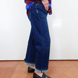 A person wearing dark blue wide-leg cropped jeans with rolled cuffs, paired with a colorful patterned blouse and black loafers, standing against a white wall on a terracotta tiled floor.