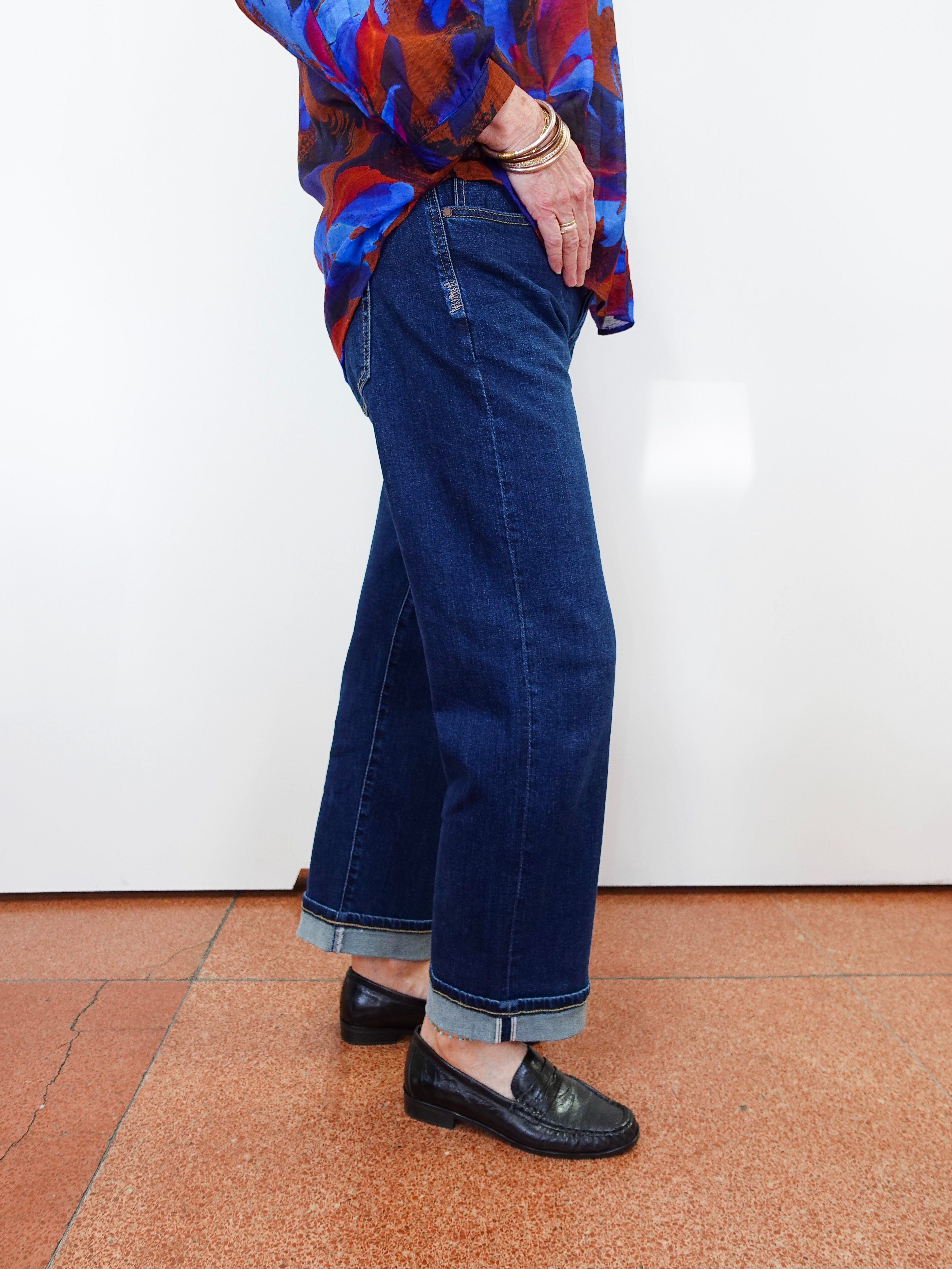 A person wearing dark blue wide-leg cropped jeans with rolled cuffs, paired with a colorful patterned blouse and black loafers, standing against a white wall on a terracotta tiled floor.