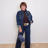 Person wearing a denim jacket and jeans holding a handbag against a white wall.