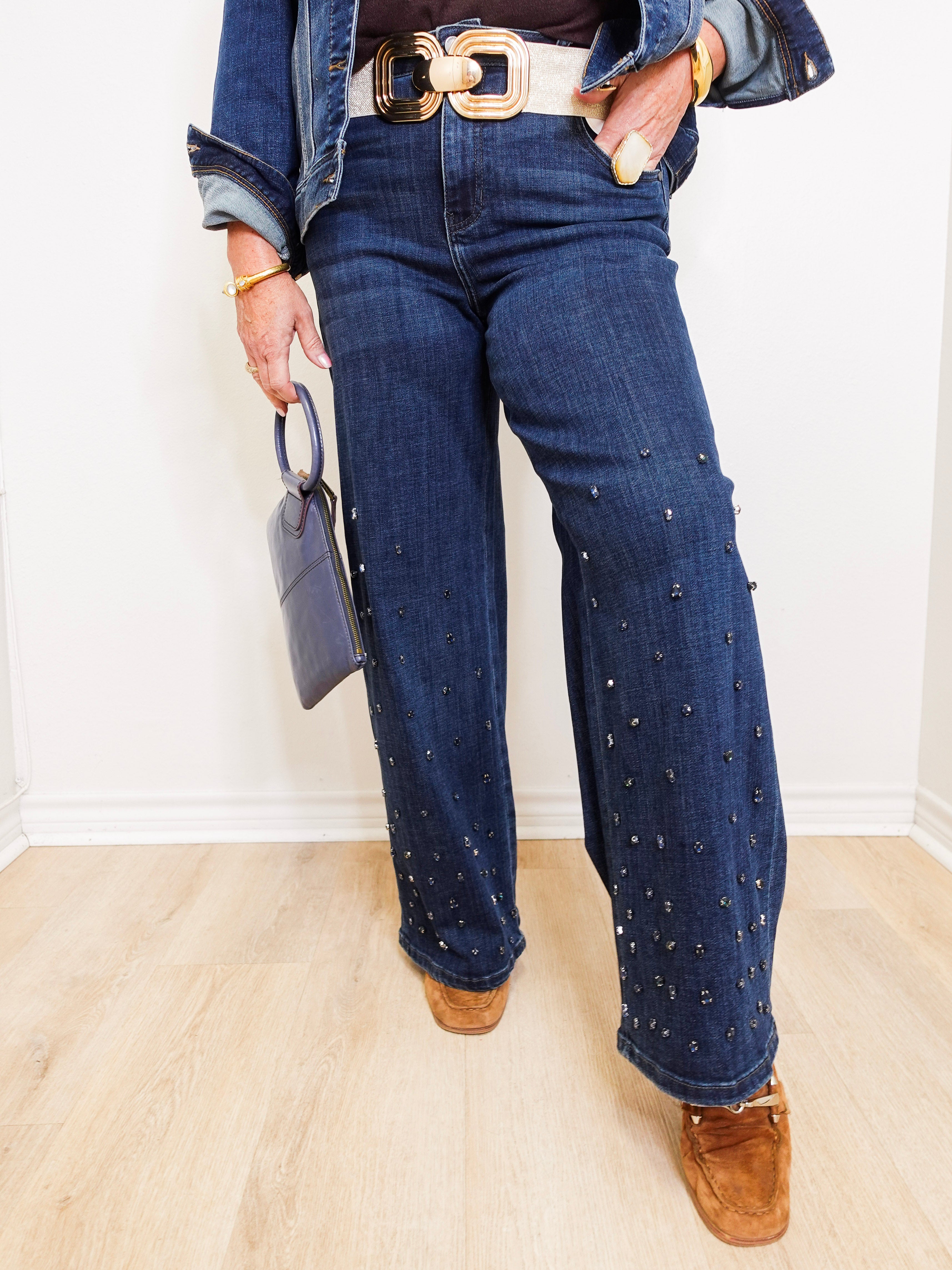 Person wearing blue jeans with decorative studs, a denim jacket, and brown shoes indoors.