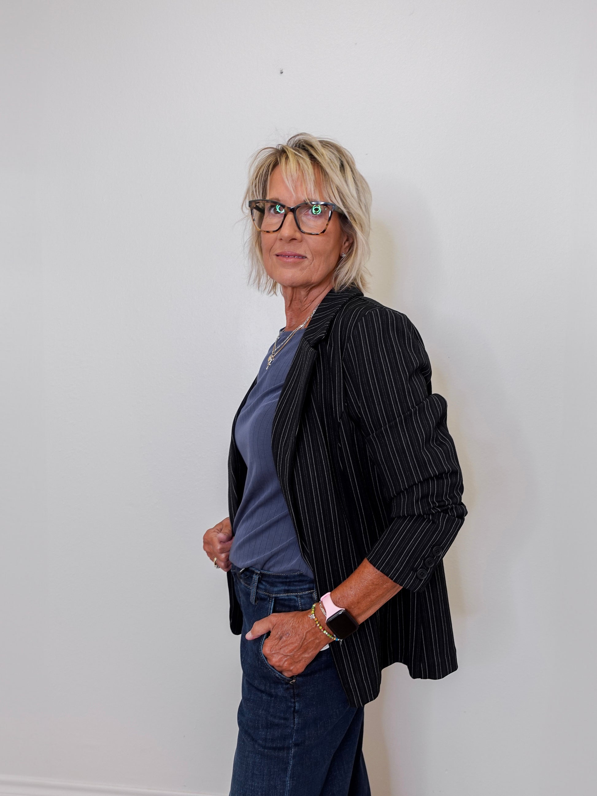 Person wearing a black pinstripe blazer over a blue shirt and jeans against a white wall.