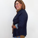A woman wearing a dark indigo denim jacket over a brown and gold patterned shirt, paired with light blue jeans, standing against a plain white background.
