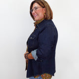 A woman wearing a dark indigo denim jacket over a brown and gold patterned shirt, paired with light blue jeans, standing against a plain white background.
