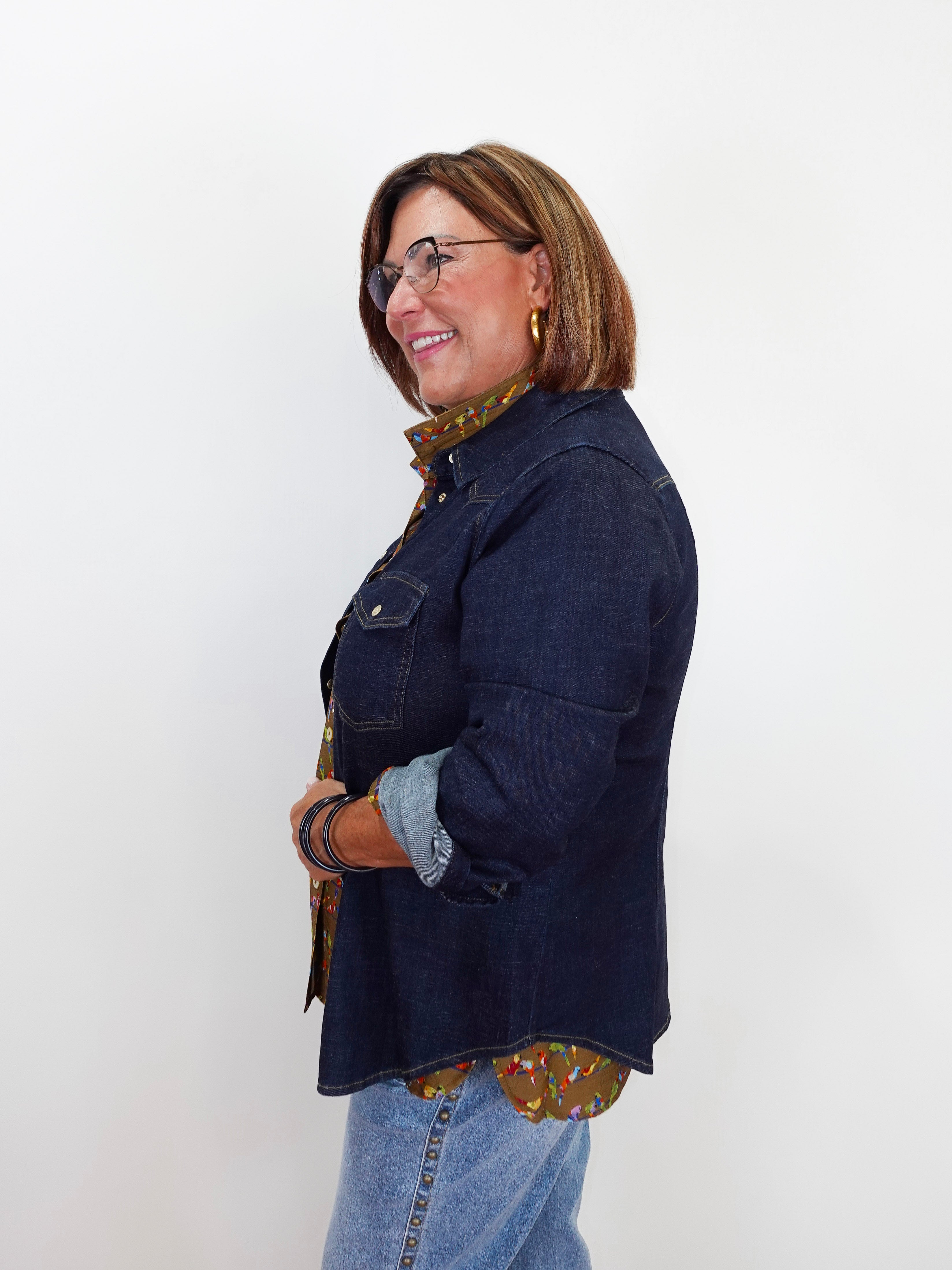 A woman wearing a dark indigo denim jacket over a brown and gold patterned shirt, paired with light blue jeans, standing against a plain white background.