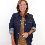 A woman wearing a dark blue denim jacket over a brown and green patterned button-up shirt, paired with light blue jeans and gold jewelry, holding a small red handbag against a plain white background.