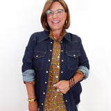 A woman wearing a dark blue denim jacket over a brown and green patterned button-up shirt, paired with light blue jeans and gold jewelry, holding a small red handbag against a plain white background.