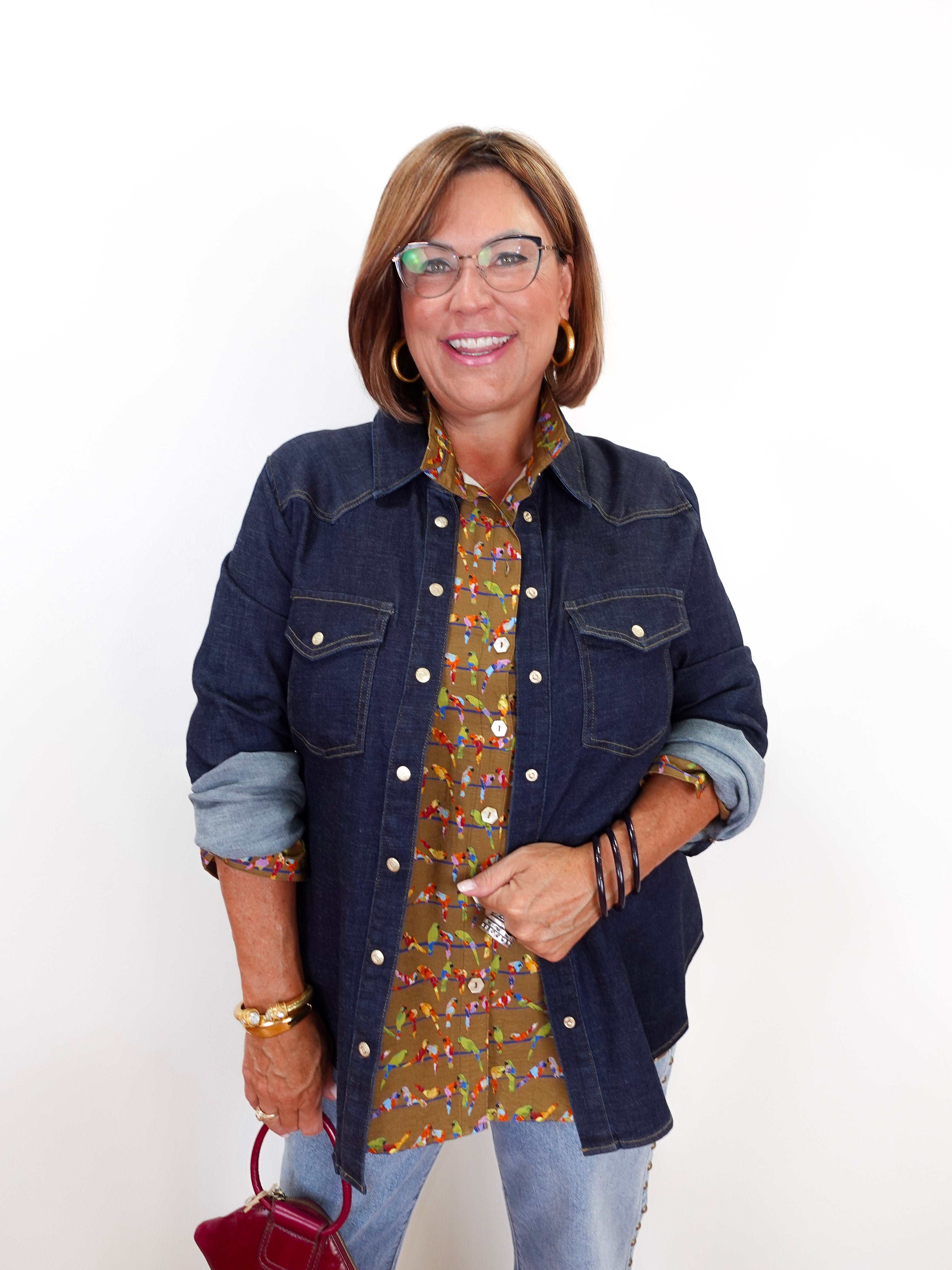 A woman wearing a dark blue denim jacket over a brown and green patterned button-up shirt, paired with light blue jeans and gold jewelry, holding a small red handbag against a plain white background.